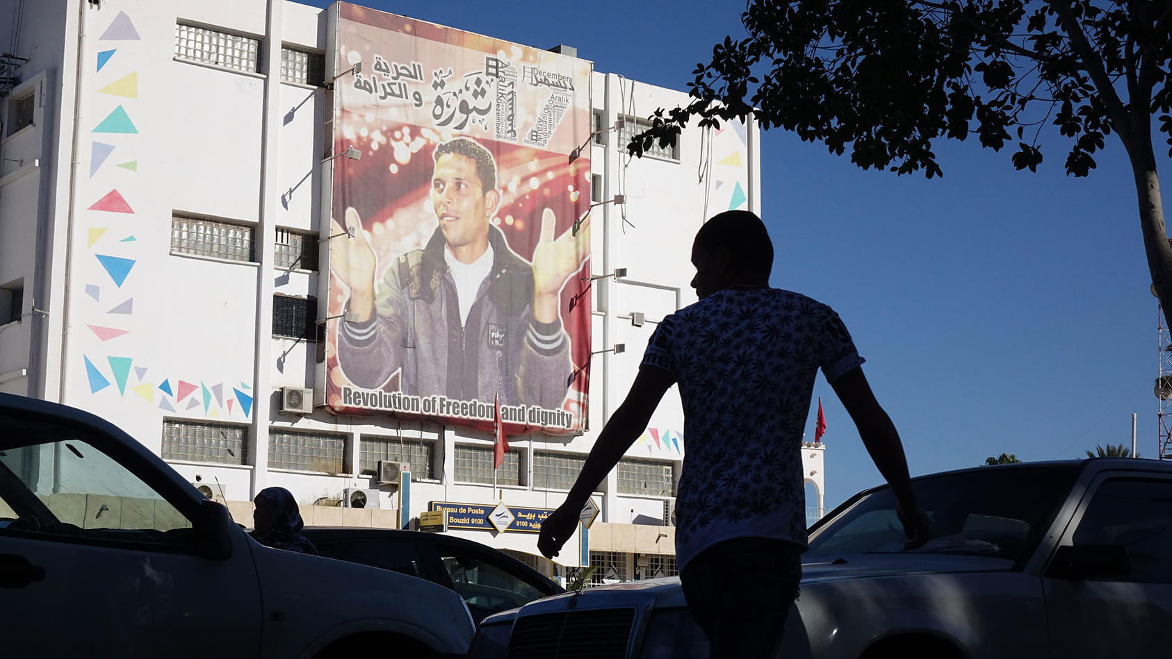 In the Tunisian city of Sidi Bouzid, cars and pedestrians bustle past the post office, with its portrait of street vendor Mohammed Bouazizi, whom Tunisians describe as a martyr to their democracy. 