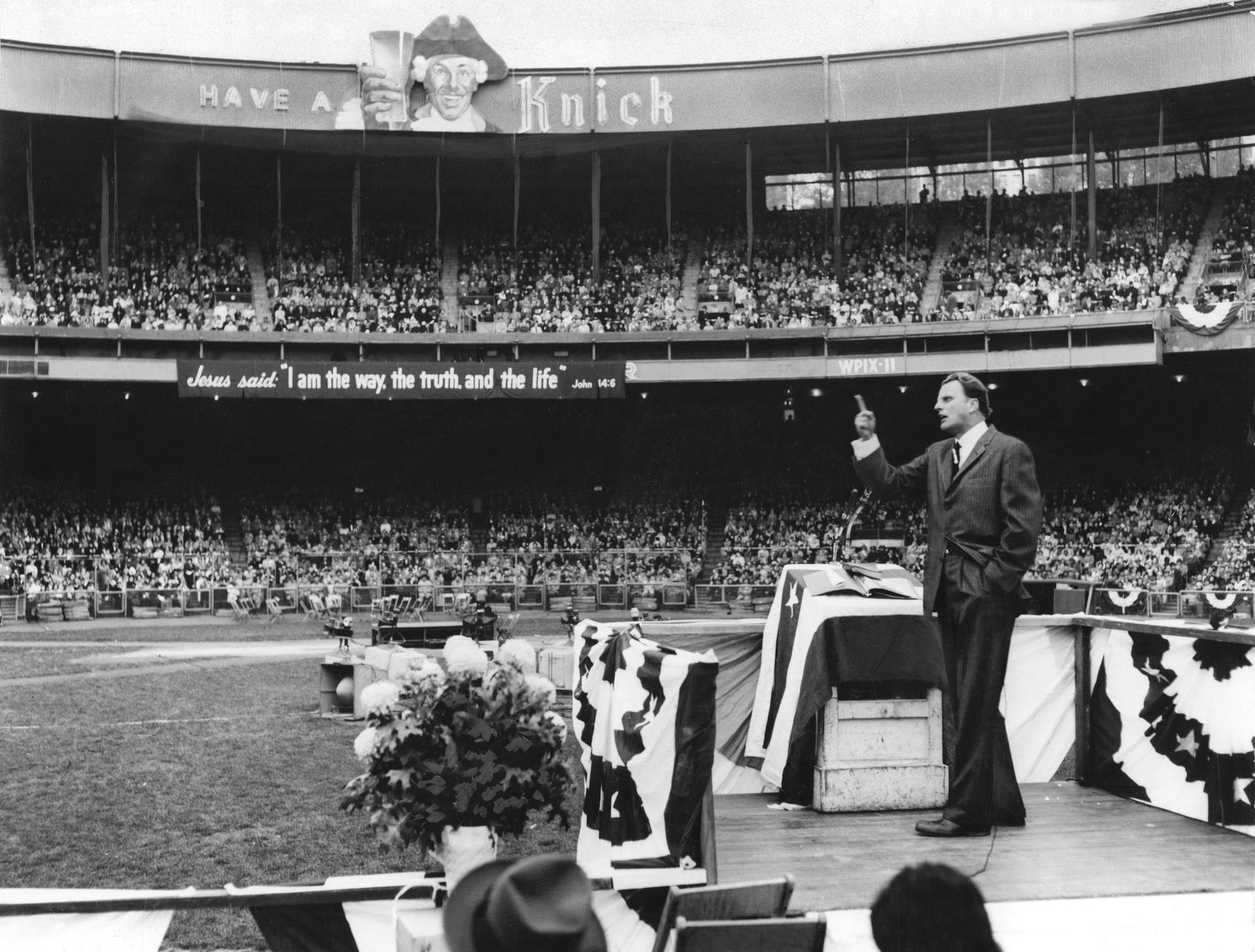 Reverend Billy Graham standing on a pulpit preaching to a stadium crowd. Photo courtesy of Allyn Baum and The New York Times